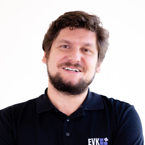 Porträt eines lächelnden Mannes mit kurzen, leicht gewellten braunen Haaren und Bart. Er trägt ein schwarzes Poloshirt mit einem „EVK“-Logo auf der Brust und steht vor einem hellen, neutralen Hintergrund.