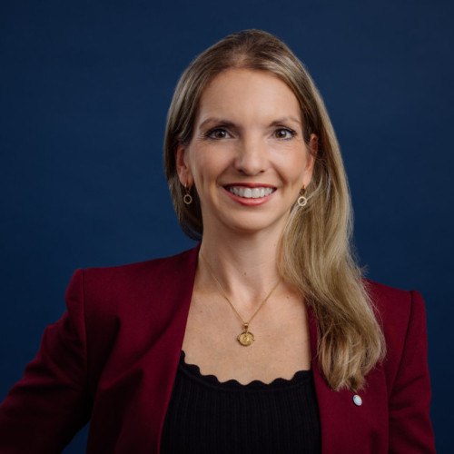 Professionelles Portrait einer Frau mit überschulterlangen blonden Haaren, die freundlich in die Kamera lächelt, vor neutral-dunkelblauem Hintergrund.