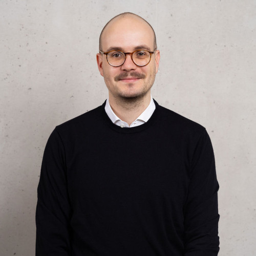 Profilbild Maximilian Eiles. Steht vor einer grauen Betonwand.Blickt freundlich lächeld in die Kamera