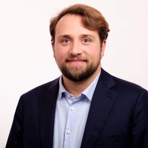 Ein Porträtfoto eines Mannes vor hellem, neutralem Hintergrund. Er schaut direkt in die Kamera und lächelt leicht. Er hat kurzes, seitlich gekämmtes braunes Haar, einen gepflegten Bart und helle Augen. Er trägt ein hellblaues Hemd mit offenem Kragen und darüber ein dunkles Sakko.