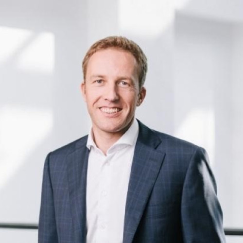 Porträt eines lächelnden Mannes mit kurzen Haaren, der einen dunkelblauen Anzug und ein weißes Hemd trägt, vor einem hellen Hintergrund mit weichem Licht.