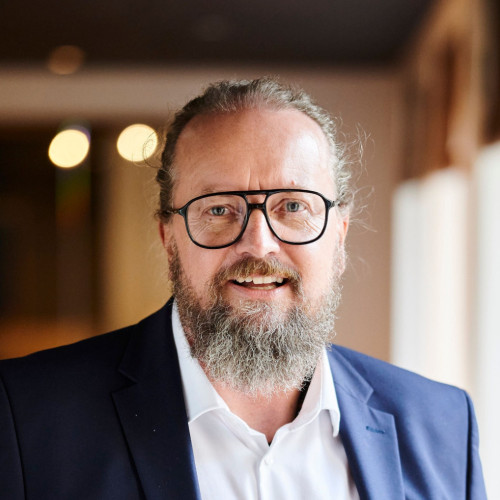 Eine Person steht frontal vor der Kamera in einem Innenraum. Sie trägt eine Brille, ein weißes Hemd und ein dunkelblaues Sakko und hat einen grauen Bart sowie nach hinten gekämmtes, graues Haar. Der Hintergrund ist weich unscharf und zeigt warme Lichtquellen sowie helle Wände.