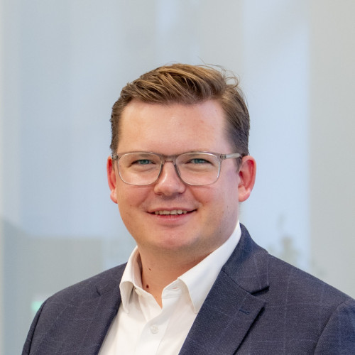 Mann mit kurzem, hellbraunem Haar trägt weißen Hemdkragen und dunkelblauen Anzug vor unscharfem Hintergrund