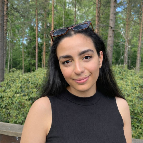 Portrait of a woman with long dark hair wearing a black top, standing outdoors in a forested area.