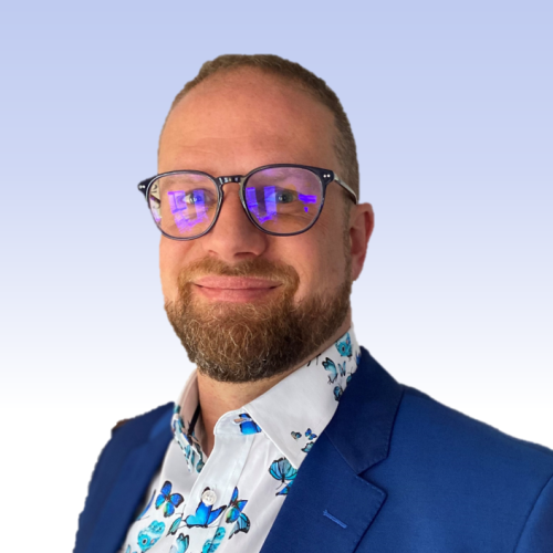 Oli is has short cropped hair and a beard. He is wearing glasses, a bright shirt and blue suit. 