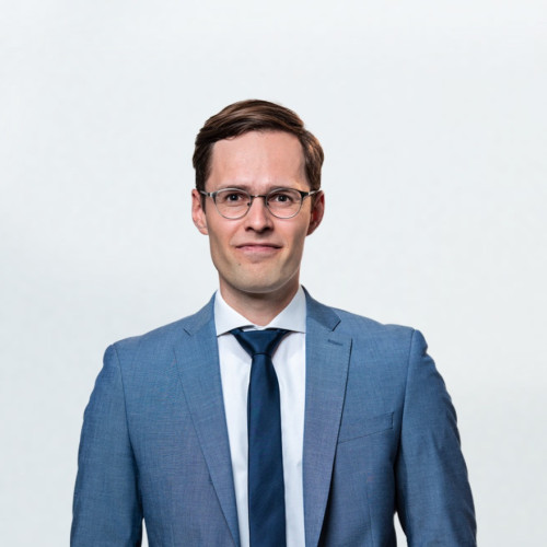 Porträt einer Person in einem blauen Anzug mit Krawatte und Brille vor hellem, einfarbigem Hintergrund.