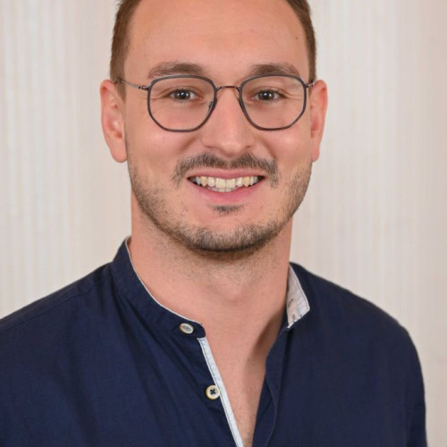 Die Aufnahme zeigt eine einzelne Person von den Schultern aufwärts in einer frontal ausgerichteten Porträtaufnahme. Die Person hat kurze, gepflegte Haare und trägt eine runde Brille mit dunklem Rand. Das Gesicht ist zur Kamera gerichtet. Bekleidet ist die Person mit einem dunkelblauen Hemd, dessen oberer Knopf geöffnet ist. Der Hintergrund besteht aus einer hellen, neutralen Fläche ohne erkennbare Gegenstände oder Text. Die Beleuchtung ist gleichmäßig und weich.