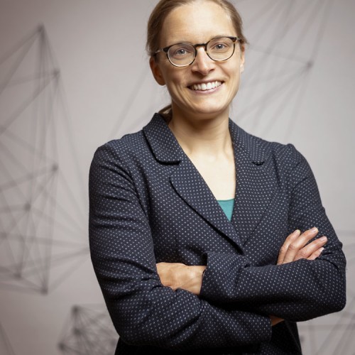 Sabine is smiling into the camera, with her arms crossed, wearing a dark blue jacket, in front of a light coloured background.