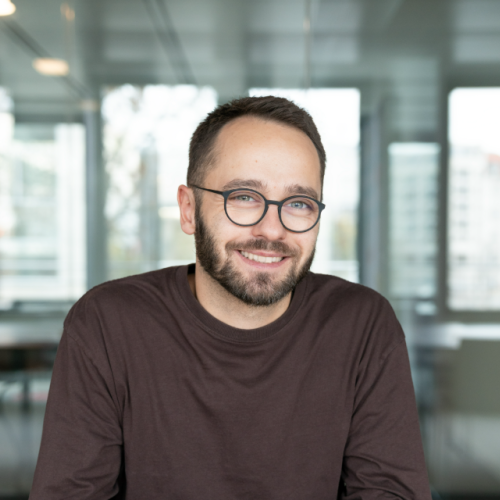 Ein lächelnder Mann mit kurzem dunklem Haar, Bart und runder Brille sitzt an einem Tisch und hat die Hände locker gefaltet. Er trägt ein langärmliges, dunkelbraunes Shirt. Im Hintergrund ist ein heller, moderner Innenraum mit großen Glasflächen und unscharf erkennbaren Gebäuden zu sehen.